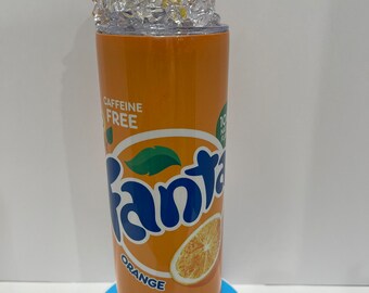 May include: Orange-colored stainless steel tumbler with a white lid and a printed image of a Fanta orange soda can. The can has the words "Fanta Orange" and "Caffeine Free" printed on it. The tumbler is filled with ice and topped with an orange slice.