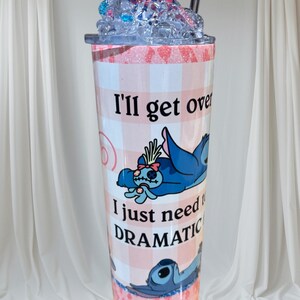 May include: A tall tumbler with a pink and white checkered pattern, featuring a blue cartoon character and the text "I'll get over it, I just need DRAMATIC." The lid is topped with blue and pink decorative elements and a straw.