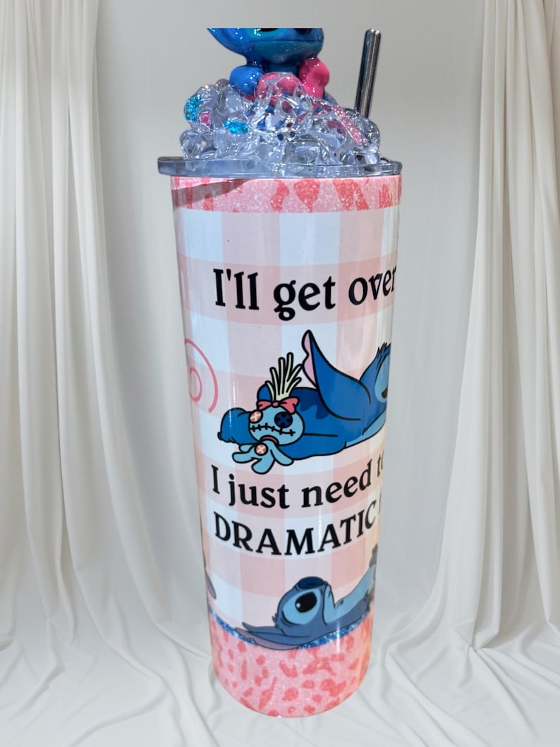 May include: A tall tumbler with a pink and white checkered pattern, featuring a blue cartoon character and the text "I'll get over it, I just need DRAMATIC." The lid is topped with blue and pink decorative elements and a straw.