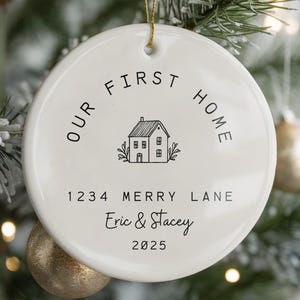 White ceramic ornament with black text that reads 'Our First Home' with a house icon and the address '1234 Merry Lane', names 'Eric & Stacey', and the year '2025'.  The ornament has a string for hanging.  The text 'Ships Fast!' is in the bottom right corner of the image.