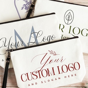 May include: Three off-white canvas zipper pouches with black wrist straps. Each pouch features different custom logo designs in blue, green, and dark red, with text including "Your Logo", "Your Custom Logo", and "And Slogan Here" printed on the front.