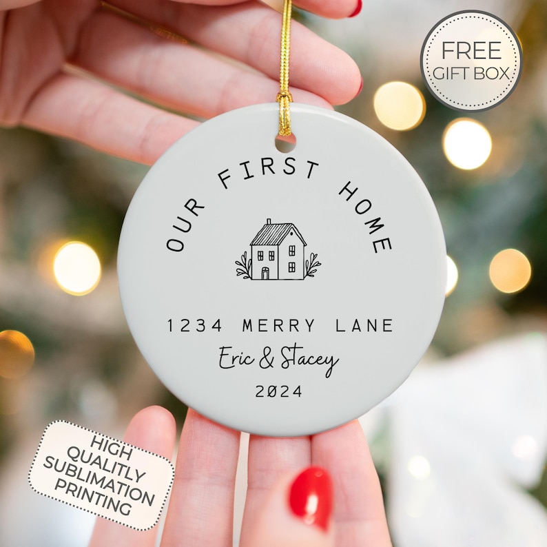 May include: White ceramic ornament with the text "Our First Home" in a black circle around a house icon. The text "1234 Merry Lane Eric & Stacey 2024" is printed below the house icon.