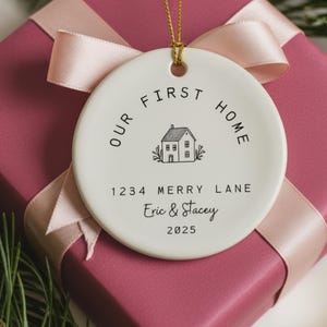White ceramic ornament with black text that reads 'Our First Home' with a house icon and the address '1234 Merry Lane', names 'Eric & Stacey', and the year '2025'.  The ornament has a string for hanging.  The text 'Ships Fast!' is in the bottom right corner of the image.