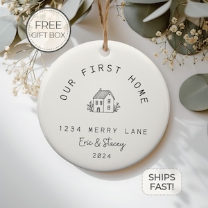 May include: White ceramic ornament with black text that reads "Our First Home" with a house illustration and the address "1234 Merry Lane" and names "Eric & Stacey" and the year "2024".