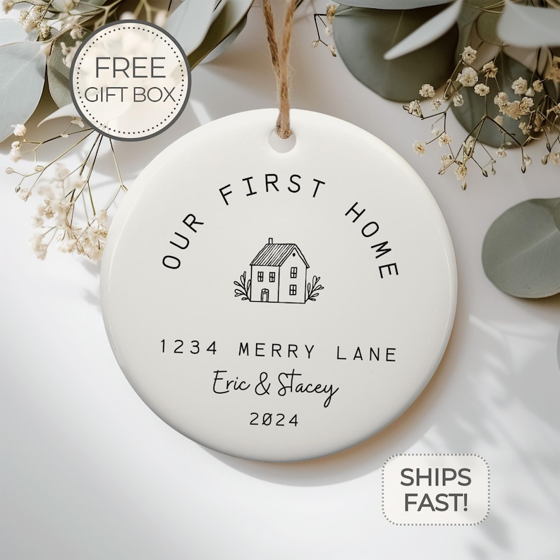 May include: White ceramic ornament with black text that reads "Our First Home" with a house illustration and the address "1234 Merry Lane" and names "Eric & Stacey" and the year "2024".
