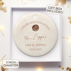May include: A white ceramic ornament with a copper-colored inscription that reads "7 Years and Counting Copper Jake & Sophia 10.07.2017". The ornament is 3 inches in diameter and comes in a gift box.