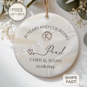 以下が含まれることがあります： 真珠をテーマにした白いセラミック製のオーナメント。"30 Years and Counting Pearl Chris & Susan 10.08.1994"と、装飾的なハートとKの文字が書かれています。