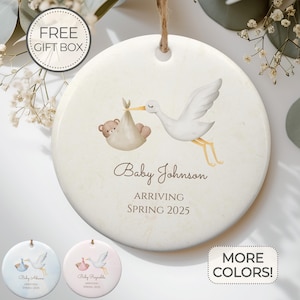 May include: A white ceramic ornament with a watercolor illustration of a stork carrying a baby in a brown teddy bear-shaped bag. The text "Baby Johnson Arriving Spring 2025" is printed below the illustration.