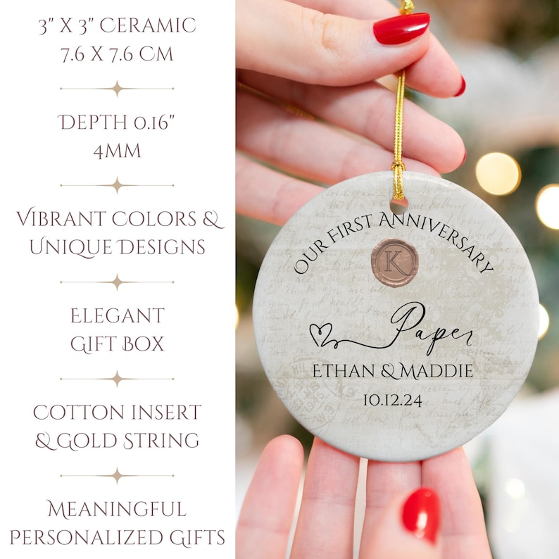 May include: A white ceramic ornament with a marbleized texture. It features the text "Our First Anniversary Paper Ethan & Maddie 10.12.24" and a gold letter "K" in a circle. The ornament is 3 inches in diameter.