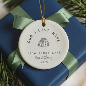 White ceramic ornament with black text that reads 'Our First Home' with a house icon and the address '1234 Merry Lane', names 'Eric & Stacey', and the year '2025'.  The ornament has a string for hanging.  The text 'Ships Fast!' is in the bottom right corner of the image.