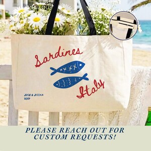 May include: A cream-colored canvas tote bag with black shoulder straps. The front features the red text "Sardines" and "Italy" with two blue fish illustrations in between. The bottom left corner displays the blue text "JOSH & JESSICA 2027".