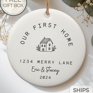 May include: White ceramic ornament with black text that reads "Our First Home" with a house illustration and the address "1234 Merry Lane" and the names "Eric & Stacey" and the year "2024".