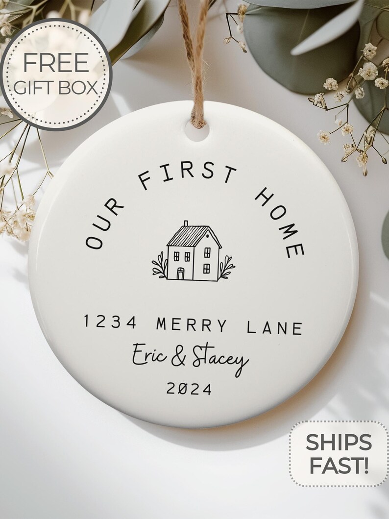 May include: White ceramic ornament with black text that reads "Our First Home" with a house illustration and the address "1234 Merry Lane" and the names "Eric & Stacey" and the year "2024".