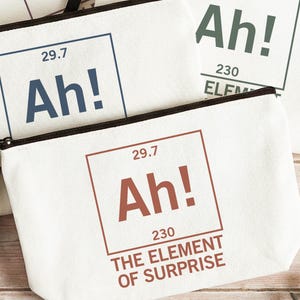 May include: Off-white canvas zipper pouches featuring a periodic table-style graphic. The design includes the text Ah!, the number 29.7, the number 230, and the phrase THE ELEMENT OF SURPRISE in blue, green, or terracotta ink with a black zipper.
