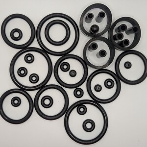 May include: A collection of black rubber O-rings and grommets of various sizes and shapes. Some have multiple holes, while others are solid. The assortment is arranged on a white surface, showcasing the different sizes and designs.