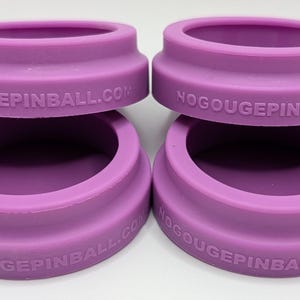 May include: Four lavender-colored, circular rubber pinball machine protectors. Each protector has a raised outer ring with the text NOGOUGEPINBALL.COM. The protectors are stacked in pairs, with one set on top of the other.