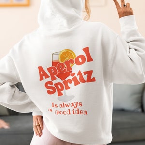 Puede incluir: Sudadera con capucha blanca con un gráfico de un vaso de Aperol Spritz y el texto "Aperol Spritz is always a good idea".