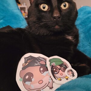 May include: A black cat with yellow eyes is the focal point of this image, with three stickers in front of it. The stickers feature cartoon illustrations of characters with tears, glasses, and hats. The cat is resting on a blue surface.