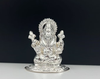 Può includere: Una statua d'argento di Ganesha con un'aureola decorativa e dettagli intricati. La statua è seduta su una base circolare.