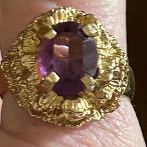 May include: A gold-toned ring featuring a large, oval-cut purple gemstone. The ring has a textured, ornate setting, giving it a vintage or antique appearance. The gemstone is faceted, reflecting light.