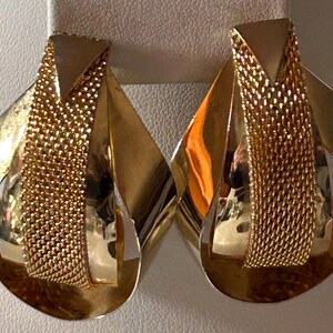 May include: A pair of gold-toned earrings with a unique design. Each earring features a combination of solid, curved metal and a central mesh-like section. The earrings have a polished finish, reflecting light.
