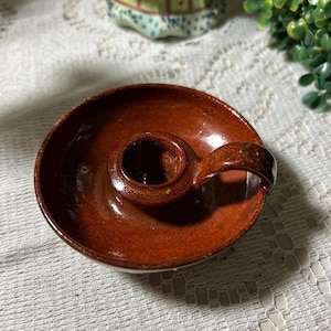 May include: A small, brown ceramic candle holder with a handle. The holder has a circular base and a raised center for holding a candle.