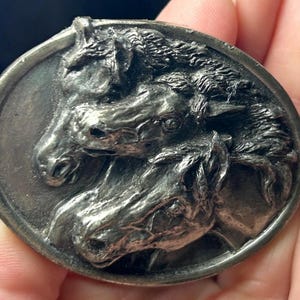 May include: A detailed, oval-shaped, silver-toned metal belt buckle featuring a relief sculpture of three horse heads. The horses are depicted with intricate detail, showcasing their manes and facial features. The buckle has a slightly aged appearance.