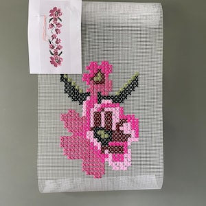 May include: A cross-stitch floral design in shades of pink, green, and brown, on a white mesh canvas. A smaller floral pattern is attached at the top. The design features a large flower with detailed petals and leaves.