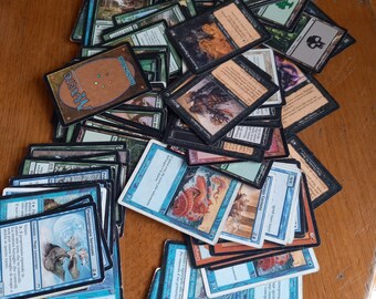 Puede incluir: Una colección de cartas de Magic: The Gathering, con una variedad de ilustraciones y tipos de cartas. Las cartas están dispuestas en una pila sobre una superficie de madera.