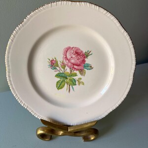 May include: A white ceramic plate with a gold rim and a pink rose design. The plate is on a gold metal stand.