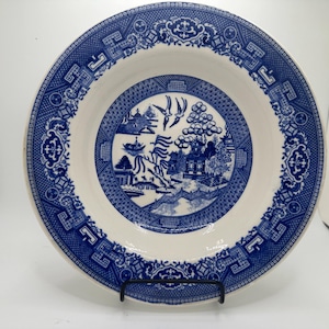 May include: A blue and white ceramic plate with a classic Willow pattern. The plate features a detailed landscape scene in the center, surrounded by a decorative border. The plate is displayed on a black metal stand, showcasing its intricate design.