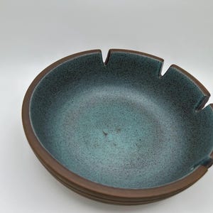 May include: A round ceramic ashtray featuring a speckled teal interior glaze and a smooth, dark brown matte rim. The circular edge includes three evenly spaced notches designed for resting items, set against a plain, neutral background.