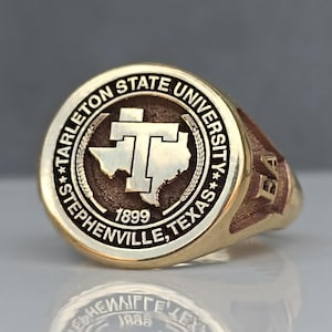May include: A gold-colored class ring with a brown and white circular design featuring the text "Tarleton State University", "Stephenville, Texas", and "1899". The ring also features a stylized outline of the state of Texas with a "T" inside.
