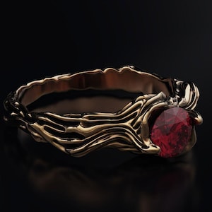 Może przedstawiać: Złoty pierścionek o teksturowanym, organicznym wzorze. Pierścionek posiada duży, okrągły, głęboko czerwony kamień szlachetny. Opaska ma płynny, przypominający gałąź wzór, tworząc unikalną i elegancką biżuterię.