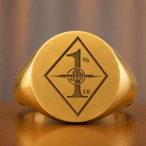 Pode incluir: Um anel de sinete dourado com uma face circular plana e uma gravação em forma de diamante. Dentro do diamante está o texto 1%ER, com um gráfico central de um punho segurando um raio, tudo com um acabamento gravado em tom escuro.