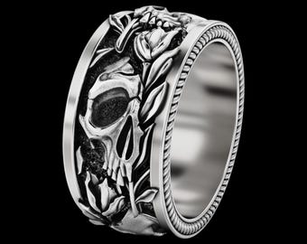 Könnte beinhalten: Ein silberner Ring mit detailliertem Schädel- und Blumendesign. Der Ring hat ein breites Band mit einem seilartigen Rand. Der Schädel ist aufwendig geschnitzt, umgeben von Blättern und Blumen, was eine gotische oder alternative Ästhetik erzeugt.