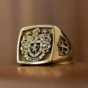 May include: A gold-toned signet ring featuring a detailed crest with a lion, a helmet, and the text ORTIZ at the base. The side of the ring displays a shield emblem with an anchor design. The metal surface has a polished finish with recessed areas.