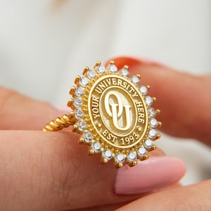 May include: A gold-colored ring featuring a university emblem with the text "YOUR UNIVERSITY HERE EST 1993". The ring has a twisted band and a halo of small, clear gemstones around the emblem.