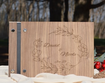 Puede incluir: Un álbum de fotos de madera con cierre de cinta gris oscuro y diseño floral grabado. El álbum está personalizado con los nombres "David & Nicole" y la fecha "Jan 25".