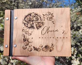 May include: A wooden recipe book with a black spine and metal accents. The cover features a laser-engraved design of a pizza with toppings, vegetables, and the text "Olivia's Recipe Book".