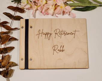 May include: A wooden photo album with the words "Happy Retirement Robb" engraved on the cover. The album is light-colored with a black spine and metal rivets. Pink and white flowers and greenery are in the background.