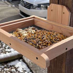 May include: A wooden bird feeder with a mesh bottom, mounted to a wooden post. The feeder is filled with a mixture of birdseed, corn, and peanuts.