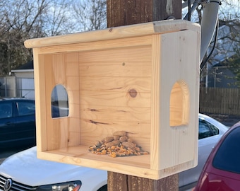 Op de afbeelding: Een houten vogelvoederhuisje met een transparante voorkant en een dak. Het voederhuisje is bevestigd aan een houten paal en is gevuld met vogelzaad. Het voederhuisje bevindt zich in een woonwijk met geparkeerde auto's op de achtergrond.