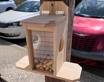 Op de afbeelding: Een houten vogelvoederhuisje met een doorzichtig plastic raam, gemonteerd op een houten paal. De voederbak is gevuld met pinda's.