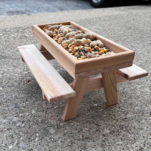 May include: A miniature wooden picnic table bird feeder filled with a mixture of birdseed, peanuts, and corn kernels.