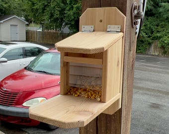 Op de afbeelding: Een houten vogelvoederhuisje met een scharnierend deksel en een platform eronder. De voederbak is gemonteerd op een houten paal en is gevuld met maïskorrels.