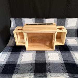 May include: A light-colored wooden box with a central compartment and two side compartments. The side compartments have circular openings. The box is made of unfinished wood and has a hinged top. The box is sitting on a black and white checkered blanket.