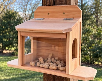 May include: A light brown wooden bird feeder with a hinged roof, a clear front panel, and arched side openings. The feeder is mounted to a dark wooden post and contains a pile of unshelled peanuts on the interior base platform.