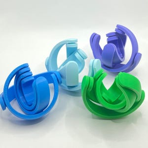May include: Four 3D-printed geometric puzzles in shades of blue, purple, and green. Each puzzle has a unique design with curved and interlocking elements. The objects are arranged on a white surface.