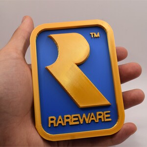 May include: A rectangular, 3D-printed plaque featuring the Rareware logo. The design includes a gold border and a gold "R" on a blue background, with the word "RAREWARE" in gold lettering below the logo. The trademark symbol is visible.